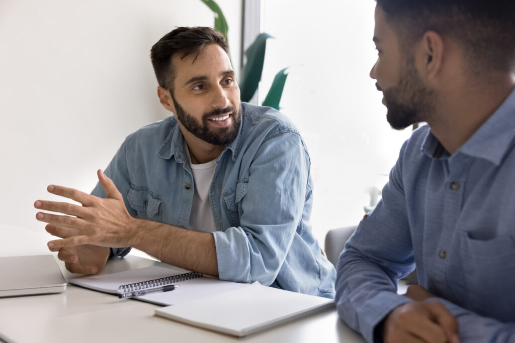 Positive handsome businessman talking to male colleague at workplace table. Diverse coworkers, two employees meeting for job discussion, cooperation on project, startup